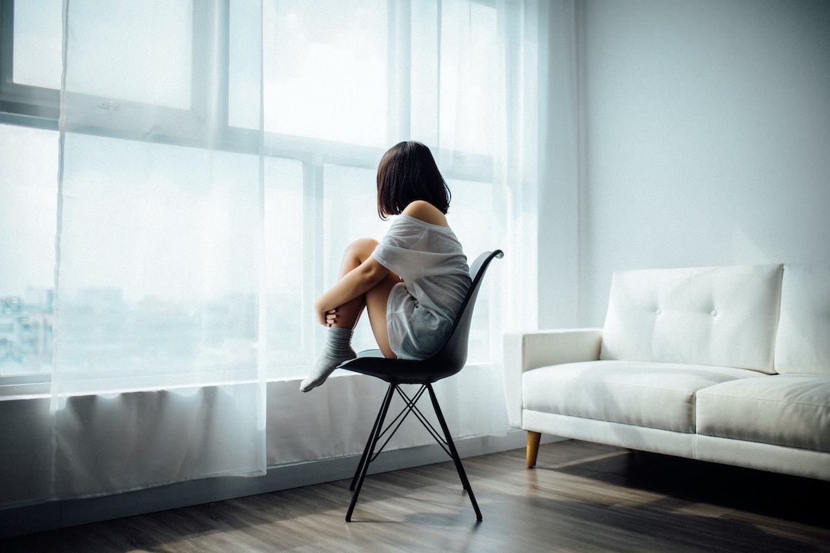 sad-woman-holding-knees-sitting-in-chair-looking-out-window